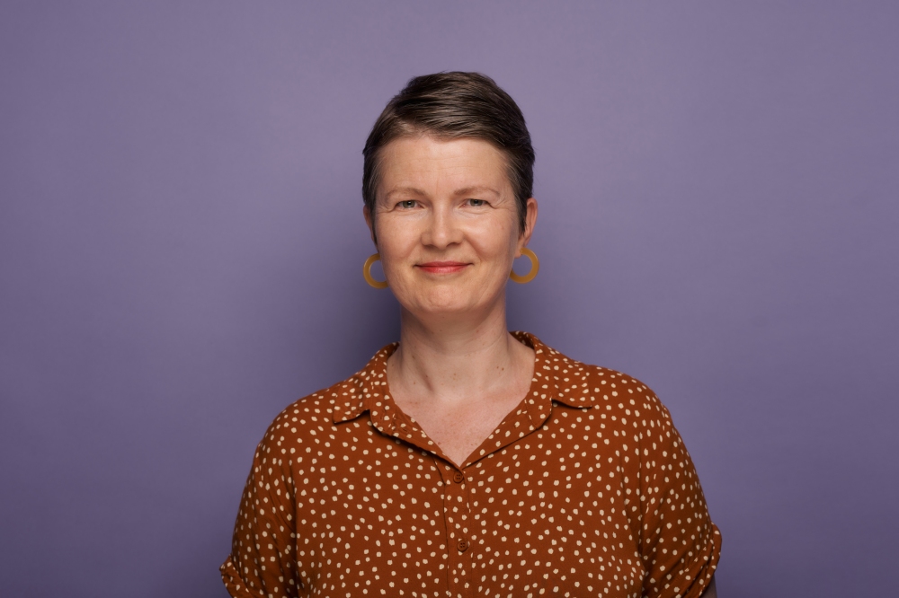 Portr?t einer l?chelnden Frau mit kurzen Haaren, brauner gepunkteter Bluse und gro?en Ohrringen vor violettem Hintergrund.