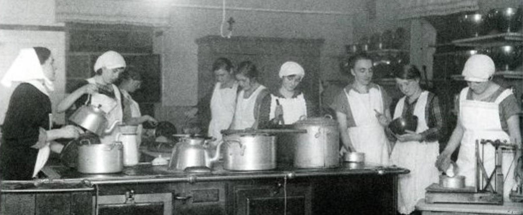 Schwarz-Weiß-Foto einer historischen Großküche mit mehreren Frauen in weißen Schürzen und Kopfbedeckungen bei der Essenszubereitung.