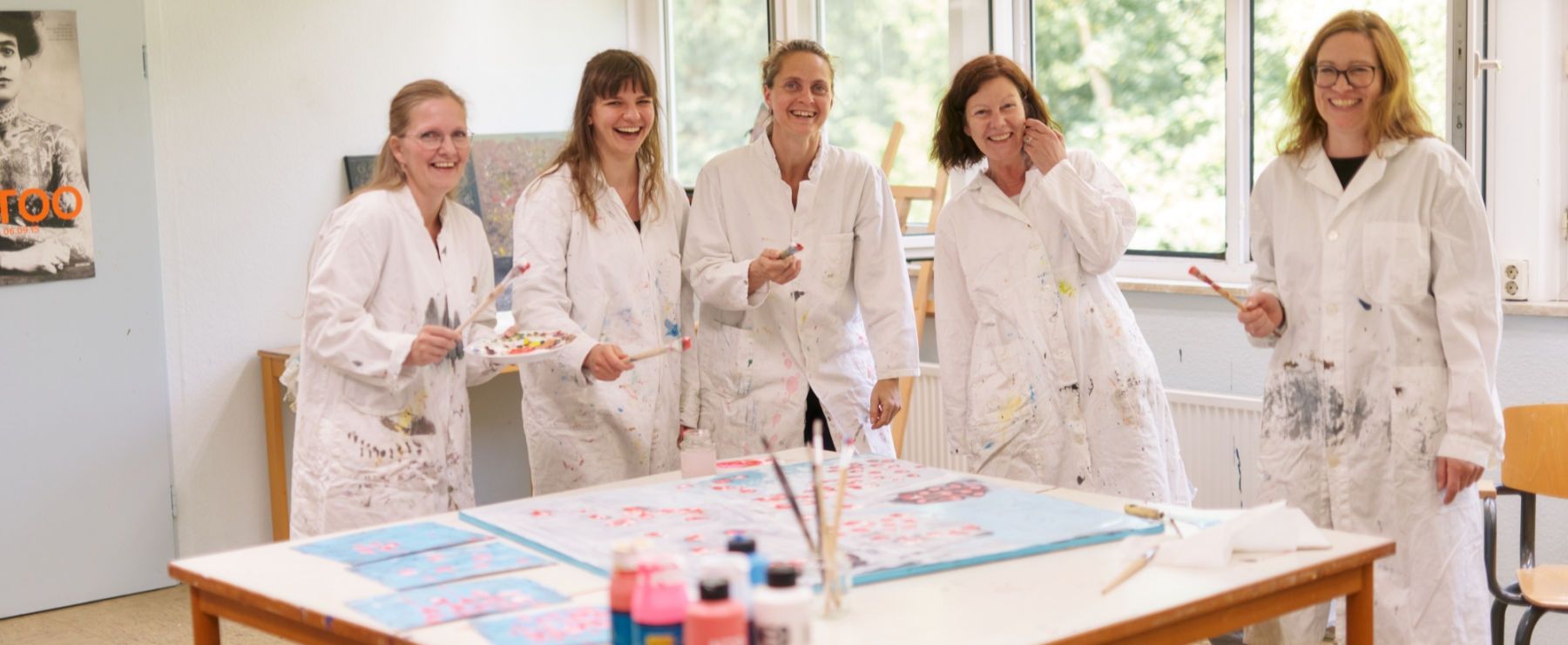 Fünf Frauen in weißen Kitteln stehen in einem Atelier. Sie halten Pinsel und Farben und malen gemeinsam. Sie lachen übers ganze Gesicht und strahlen viel Fröhlichkeit aus.