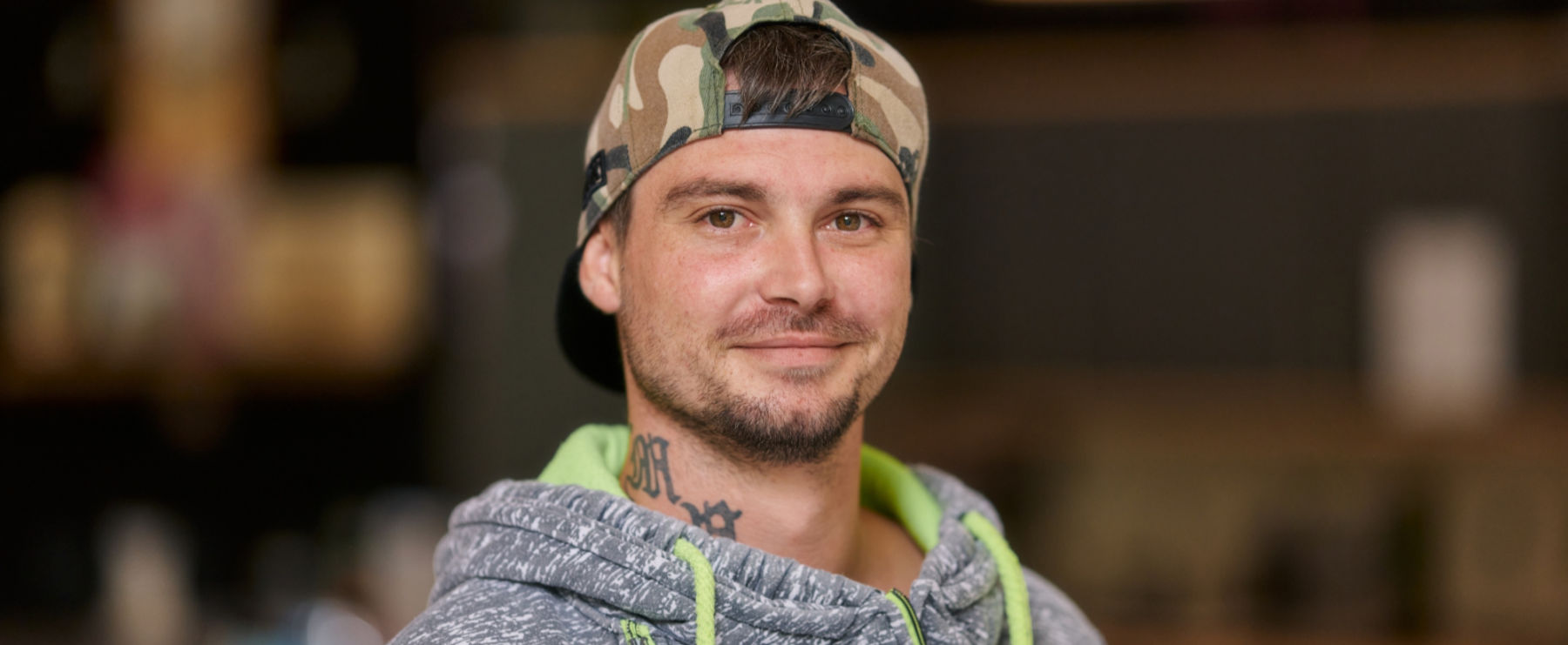 Junger Mann mit Bart, Camouflage-Cap und grauem Hoodie mit grünen Akzenten lächelt freundlich in die Kamera. Unscharfer Hintergrund mit warmem Licht.