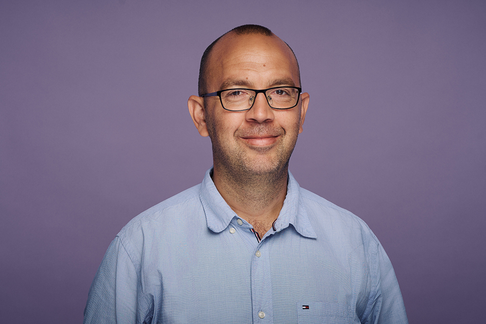 Portr?t eines Mannes mit Brille und blauem Hemd vor einem violetten Hintergrund. Er l?chelt freundlich in die Kamera.
