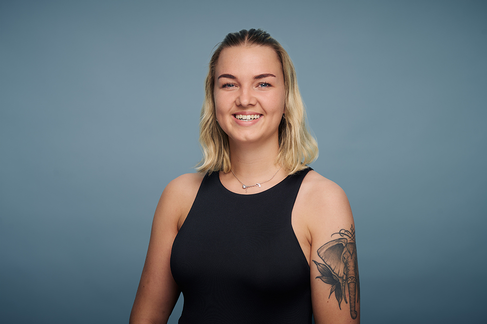 Porträt einer lächelnden jungen Frau mit schulterlangem blondem Haar vor blauem Hintergrund. Sie trägt ein schwarzes Top, eine Perlenkette und hat ein Elefantentattoo am Oberarm.