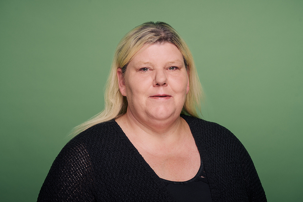 Porträt einer Frau mit schulterlangem blondem Haar vor einem grünen Hintergrund. Sie trägt ein schwarzes Oberteil.