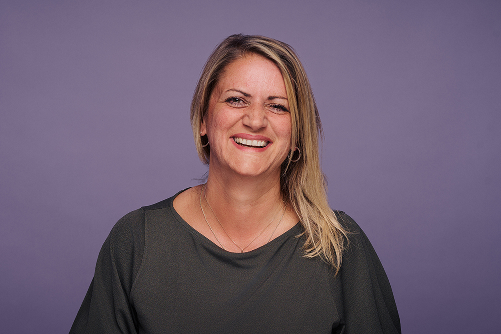 Porträt einer lächelnden Frau mit schulterlangem blondem Haar vor einem violetten Hintergrund. Sie trägt ein dunkelgrünes Oberteil.