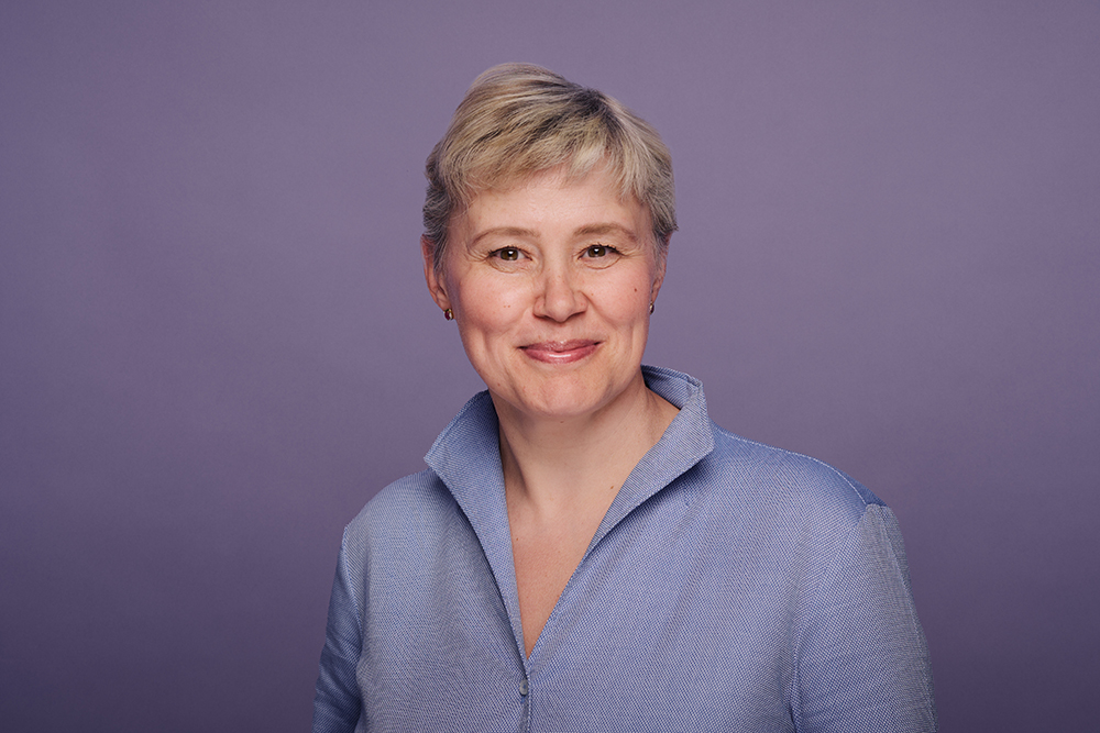 Porträt einer lächelnden Frau mit kurzem, blondem Haar vor einem violetten Hintergrund. Sie trägt eine hellblaue Bluse mit Stehkragen.