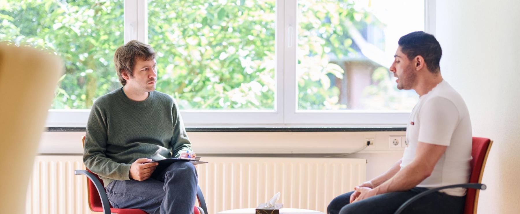 Zwei M?nner sitzen vor einem sonnigen Fenster auf roten St?hlen und f?hren ein Gespr?ch. Einer h?lt ein Klemmbrett mit Notizen.