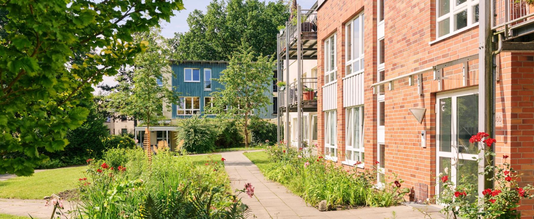 Rote Backstein- und blaue Holzgeb?ude in einer gepflegten Gartenanlage mit Blumen und gr?nen B?umen an einem sonnigen Tag.