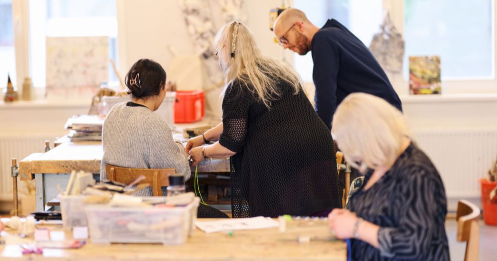 3 Personen arbeiten gemeinsam an einem kreativen Projekt in einer Werkstatt. Im Vordergrund sitzt eine blonde Frau an einem Tisch und arbeitet an einem Projekt.