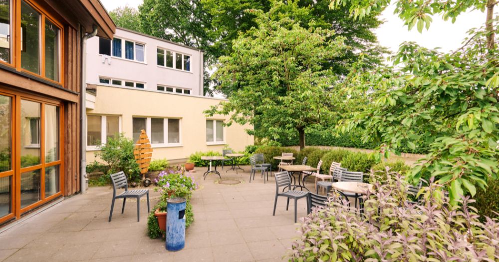 Eine Terrasse im Gr?nen mit Sitzgelegenheiten und Tischen vor einem modernen Geb?ude mit gro?en Fenstern.