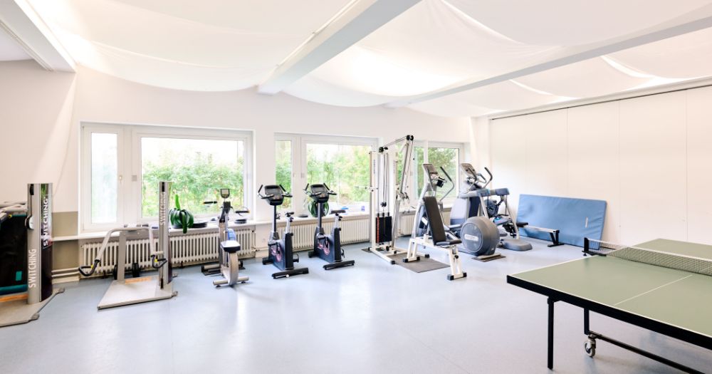 Heller Fitnessraum mit verschiedenen Trainingsger?ten, darunter Fahrradergometer, Kraftmaschinen und eine Tischtennisplatte.