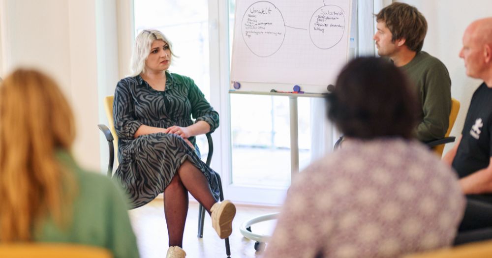 Gruppengespr?ch in einem Raum mit Flipchart. Eine hellblonde Frau in gemustertem Kleid spricht, w?hrend die anderen Personen zuh?ren.