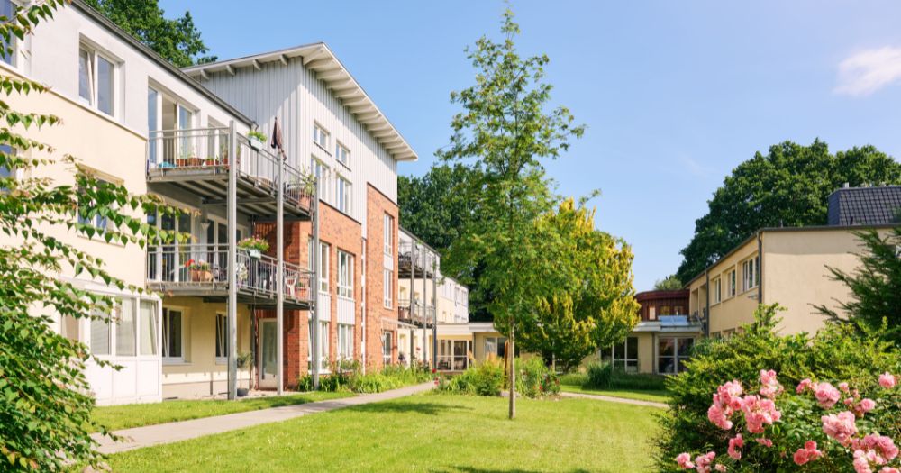 Moderne Backsteingebäude mit Balkonen, umgeben von einer grünen Gartenanlage mit blühenden Rosen, Bäumen und gepflegten Wegen.
