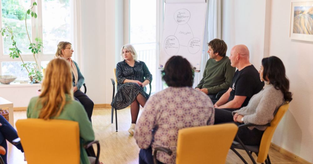 Gruppe von Menschen sitzt im Stuhlkreis in einem hellen Raum. Eine Frau spricht, hinter ihr ein Flipchart mit Notizen.