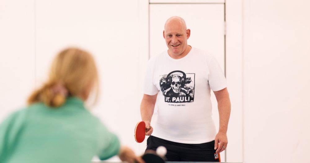 Mann im weißen T-Shirt spielt Tischtennis mit einer Frau in grünem Pullover. Er hält einen Schläger und lächelt, während er dem fliegenden Ball nachschaut.