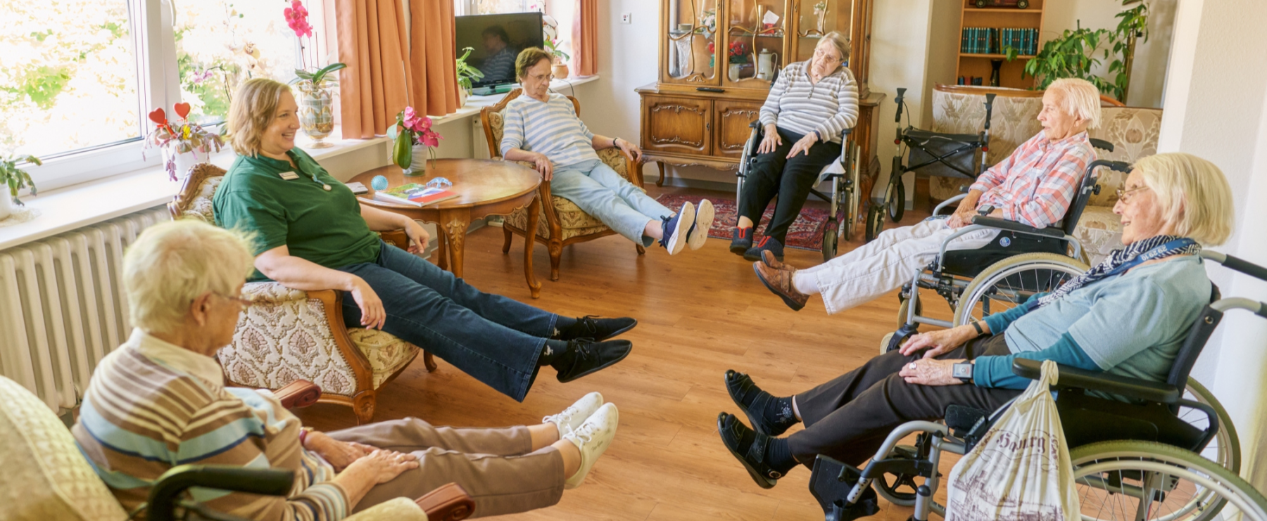 Senior:innen und eine Betreuerin sitzen in einem gemütlichen Raum und machen gemeinsam eine Gymnastikübung mit gestreckten Beinen.