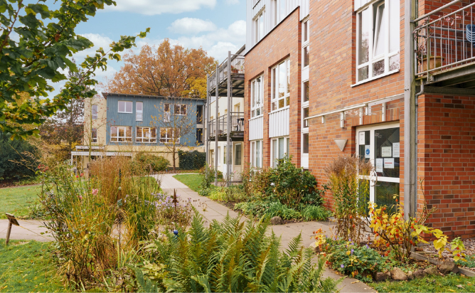 Moderne Backstein- und Holzgeb?ude mit gro?en Fenstern. Sie sind umgeben von gr?nen Beeten, durch die Gehwege zu den Geb?uden f?hren.