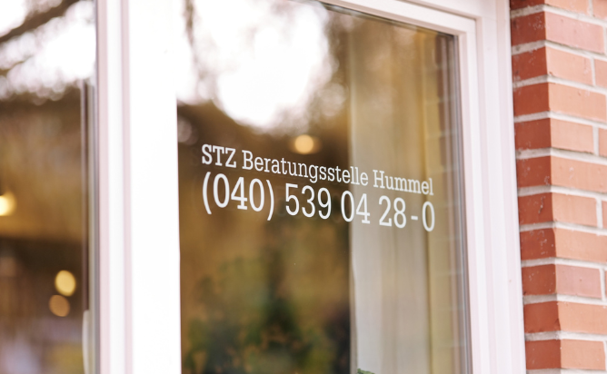 Eine Fensterscheibe mit weißer Schrift, die die Aufschrift 'STZ Beratungsstelle Hummel' und eine Telefonnummer (040) 539 04 28-0 zeigt. Eine Spiegelung auf dem Glas zeigt einen verschwommenen Hintergrund mit Bäumen und Licht.