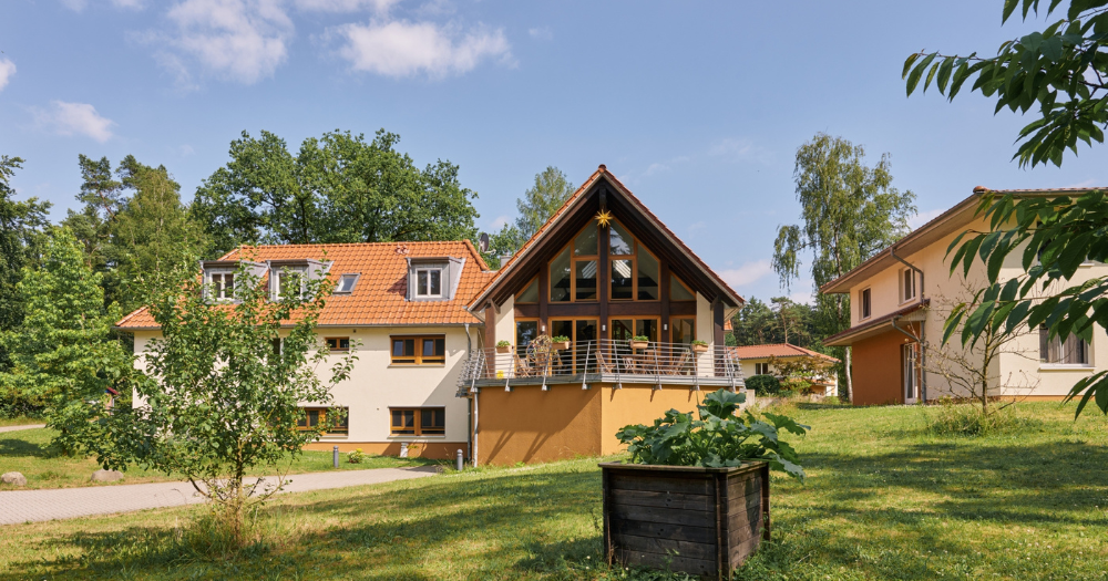 Mehrere helle H?user mit roten D?chern liegen verstreut um eine Wiese mit Hochbeet. Eines hat eine gro?e Fensterfront und eine Terrasse.