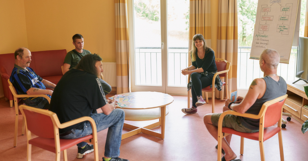 Gruppe von f?nf Menschen sitzt in einem Stuhlkreis in einem hellen Raum mit rotem Fu?boden. Eine Frau neben einem Flipchart moderiert das Gespr?ch.