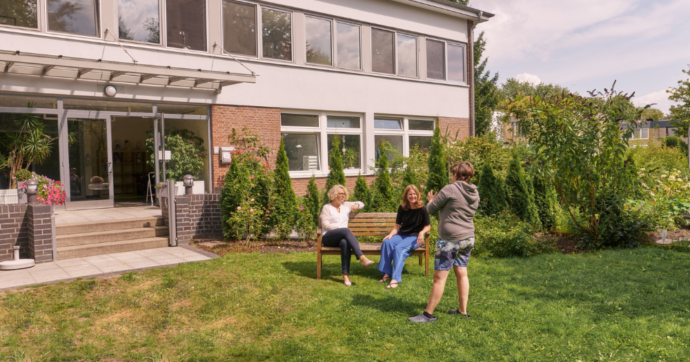 Zwei Frauen sitzen auf einer Gartenbank und unterhalten sich mit einer anderen Frau, die vor ihnen steht. Im Hintergrund ist ein modernen Gebäude zu erkennen.