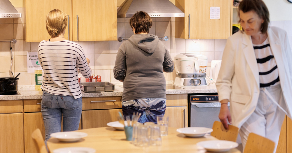 Drei Frauen in einer Gemeinschaftsküche: Zwei bereiten Essen zu, eine in weißem Anzug wirft einen prüfenden Blick auf den gedeckten Tisch.