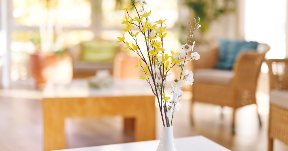 Detailaufnahme einer wei?en Vase mit gelben und wei?en Bl?tenzweigen auf einem Tisch. Im Hintergrund gem?tliche Rattansessel. Warme Atmosph?re.