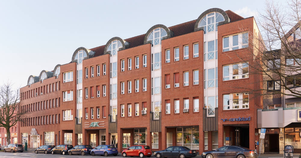 Modernes, mehrst?ckiges Backsteingeb?ude mit gro?en Fenstern. Autos parken davor, und verschiedene Firmenlogos sind sichtbar.