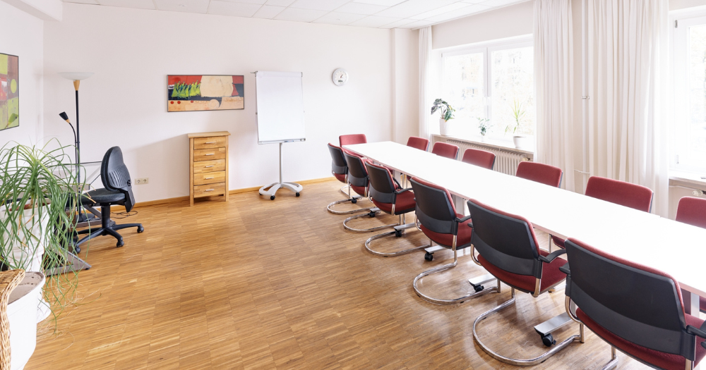 Heller Konferenzraum mit langem Tisch, roten St?hlen, Flipchart, Pflanzen und Fenstern mit wei?en Vorh?ngen.