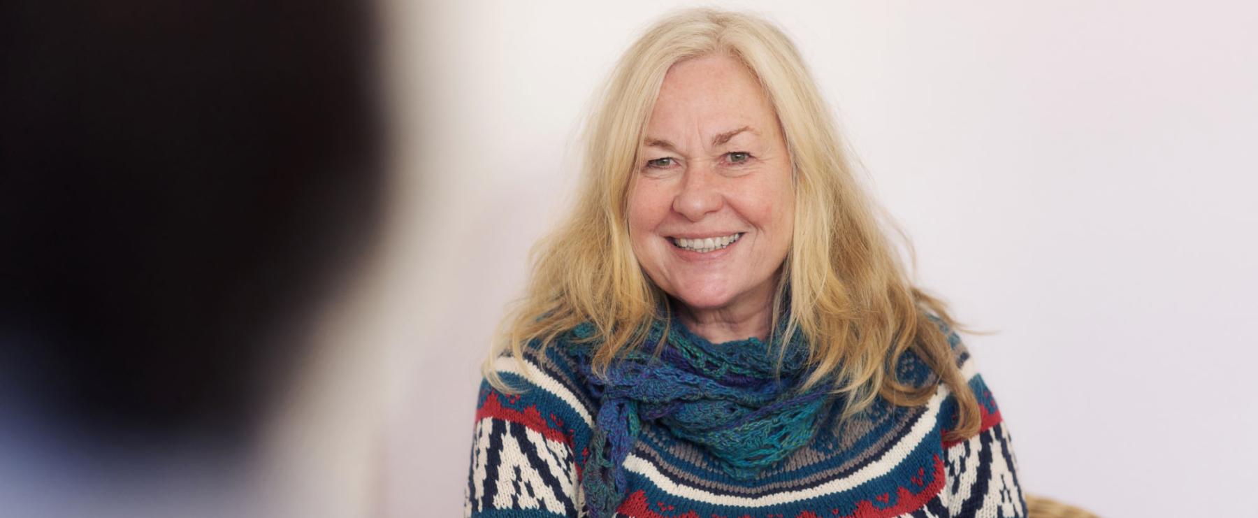 Lächelnde Frau mit blonden Haaren, bunter Strickjacke und Schal sitzt entspannt vor einem hellen Hintergrund.