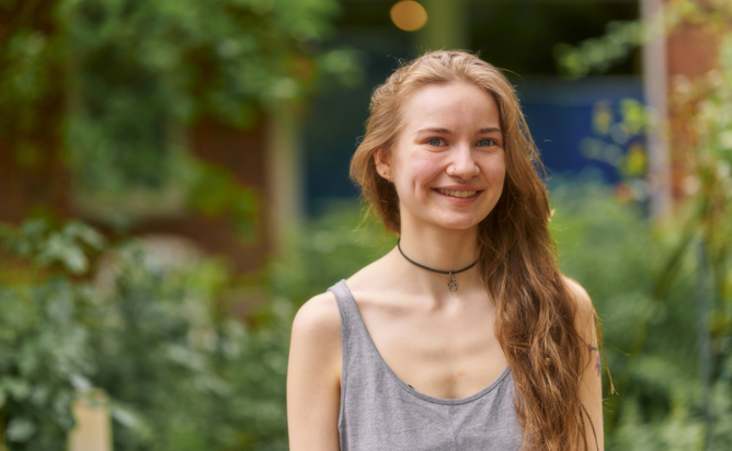 Junge Frau mit langen Haaren und grauem Top lächelt im Grünen, im Hintergrund sind Bäume und ein Gebäude sichtbar.