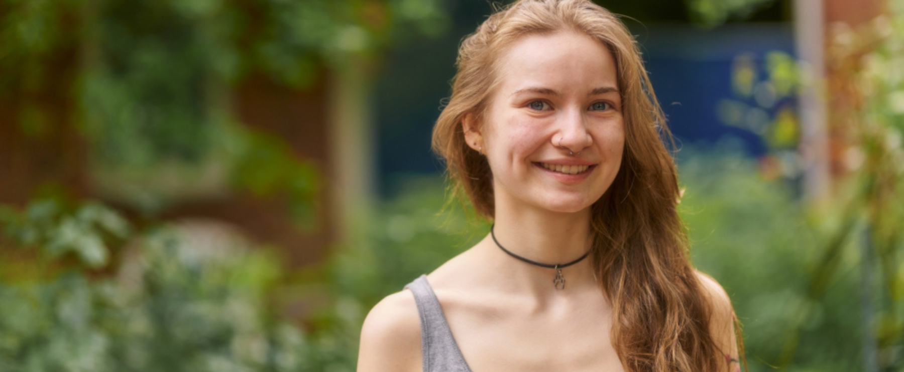 Junge Frau mit langen Haaren und grauem Top steht im Grünen und lächelt in die Kamera. Im Hintergrund sind Bäume und ein Gebäude sichtbar.