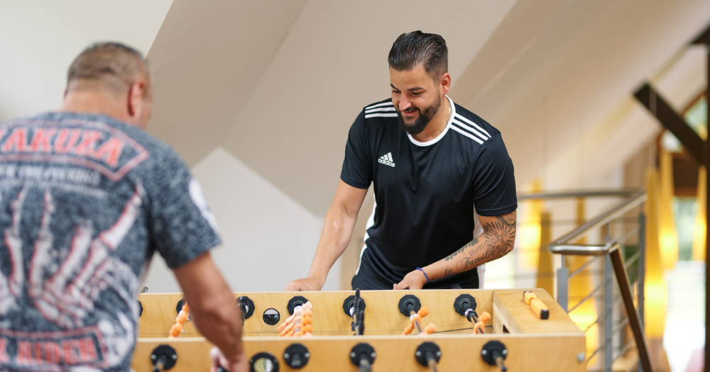Zwei M?nner spielen Tischkicker. Einer tr?gt ein schwarzes Adidas-Shirt, der andere ein gemustertes T-Shirt mit roter Aufschrift.