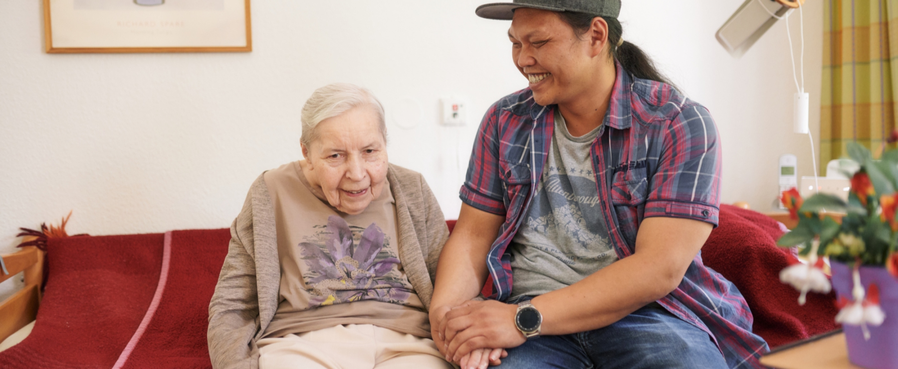 Seniorin und Pflegekraft sitzen im Wohnbereich des Seniorenzentrums St. Markus gemeinsam auf einem Sofa.