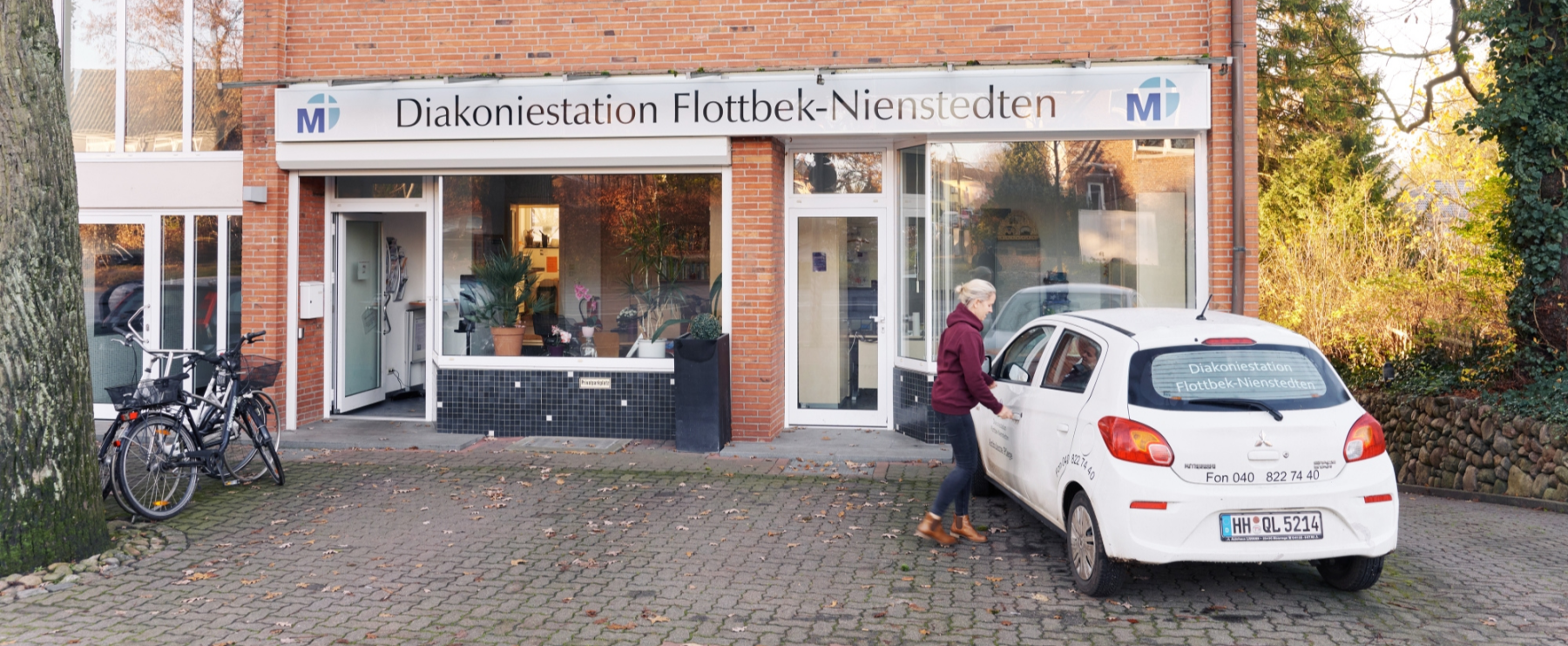 Auffahrt der Ambulanten Pflege St. Markus, hinter einer sch?nen gr?nen Hecke stehen eine Mitarbeiterin und ein Firmenfahrzeug.
