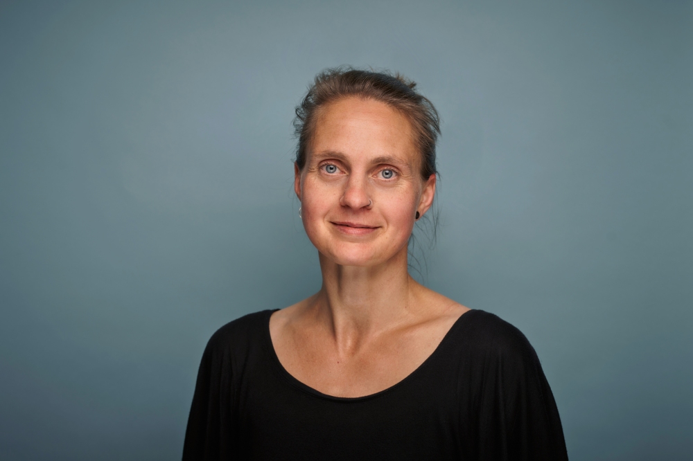 Portr?t einer Frau mit zur?ckgebundenem Haar und Nasenpiercing vor blau-grauem Hintergrund. Sie l?chelt leise in die Kamera.