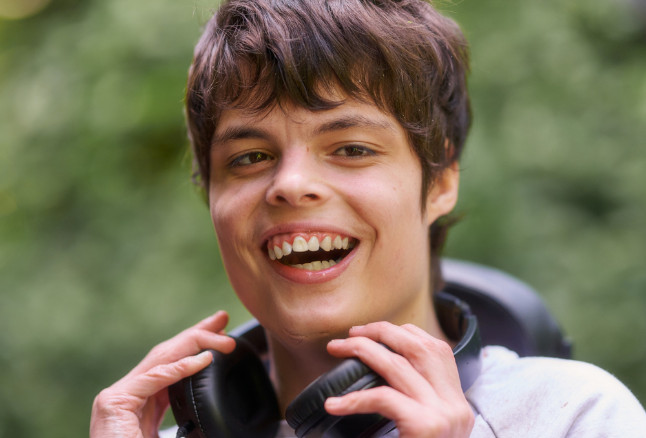 Ein junger Mensch mit braunen Haaren und Kopfhörern um den Hals lacht fröhlich in die Kamera. Der Hintergrund ist grün und verschwommen.