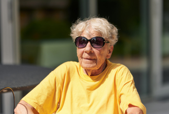 Eine ältere Frau mit kurzen grauen Haaren und Sonnenbrille sitzt entspannt in der Sonne. Sie trägt ein gelbes T-Shirt und blickt nach vorne in die Kamera.'