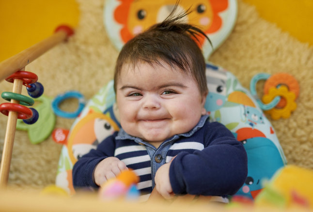 Ein fröhliches Baby mit dunklen Haaren sitzt in einer bunten Babywippe mit Spielbogen. Es ist umgeben von weichen Stofftieren und Spielzeug.