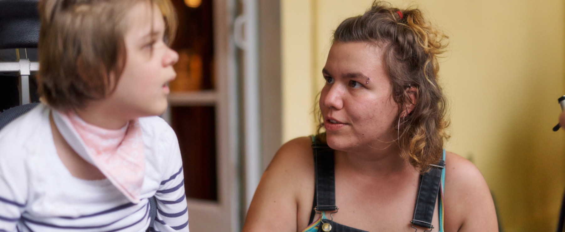 Eine junge Frau mit lockigem Haar spricht mit einem Kind mit Halstuch und gestreiftem Pullover. Sie lächelt und trägt eine Latzhose. Die Begegnung wirkt innig.