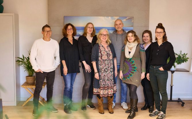 Teamfoto mit acht Personen in einem freundlich gestalteten Raum. Sie stehen vor einem Wandbild mit Meerblick und lächeln direkt in die Kamera.
