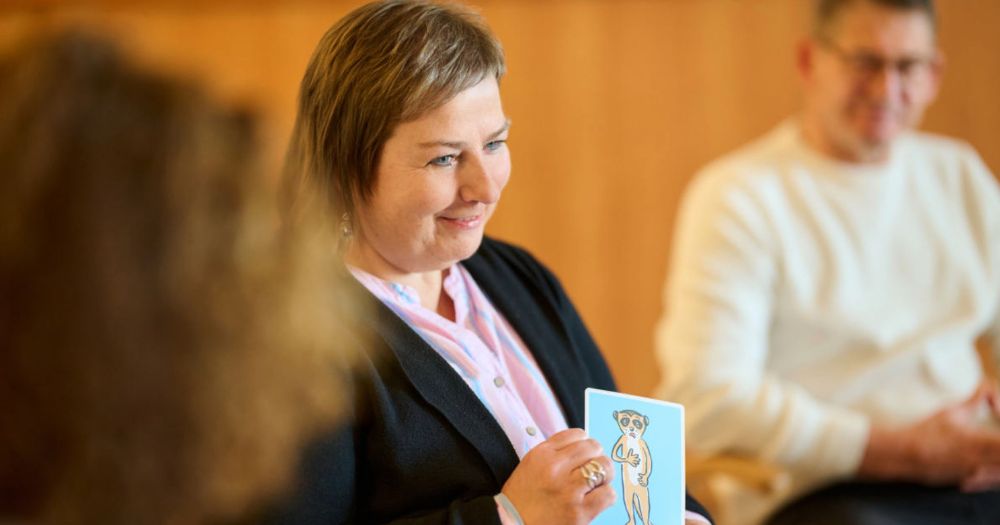 Eine Frau h?lt eine Karte mit einem gezeichneten Erdm?nnchen und l?chelt still. Im Hintergrund sind verschwommen weitere Personen wie in einem Stuhlkreis zu sehen.