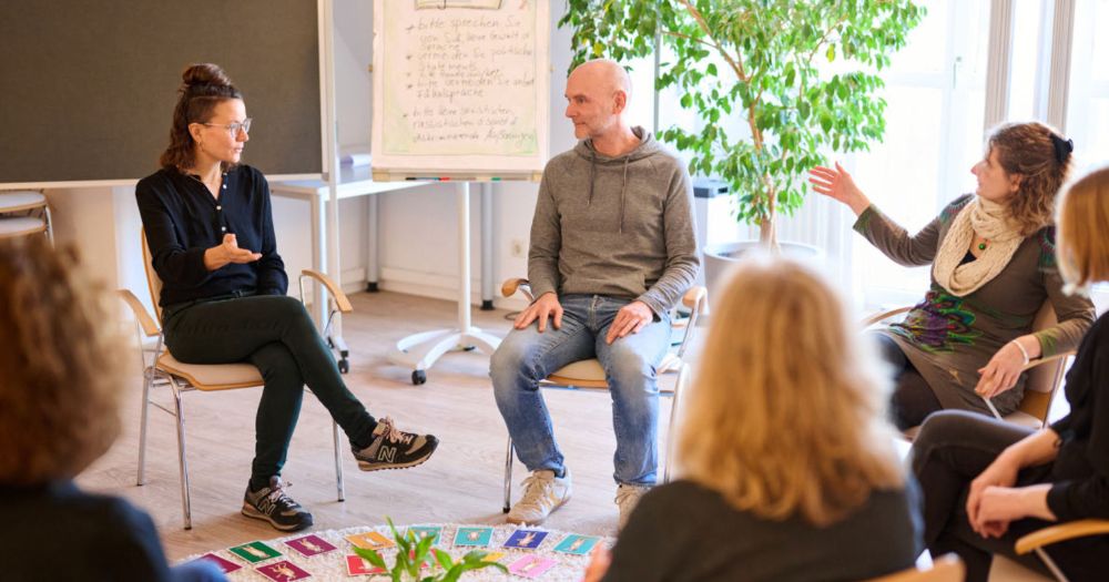 Eine Gruppe sitzt in einem Stuhlkreis und diskutiert. Eine Frau spricht, w?hrend die anderen zuh?ren. Flipchart und Pflanzen im Hintergrund.