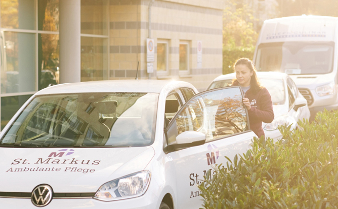 Auto des Pflegedienstes St. Markus mit Mitarbeiterin vor dem Seniorenzentrum bei Sonnenschein.