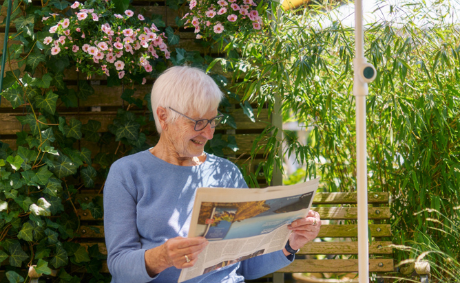 Eine ältere Frau mit Brille sitzt lächelnd auf einer Holzbank. Sie liest Zeitung und ist von blühenden Blumen und grünen Pflanzen umgeben.