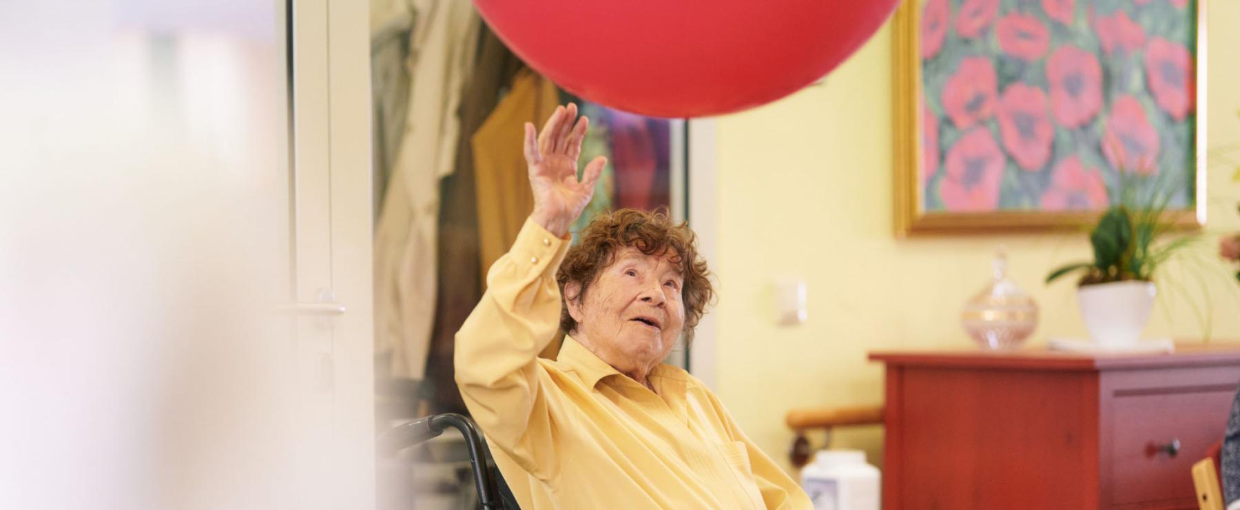 ?ltere Dame im Rollstuhl spielt mit einem gro?en roten Ball in der Luft. Sie befindet sich in einem freundlich gestalteten Raum mit Blumenbild und Pflanzen.