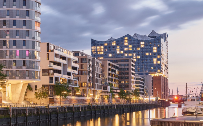Stimmungsvolle Abendaufnahme von der HafenCity mit moderner Wohnanlage links von der beleuchteten Elbphilharmonie. Im Vordergrund spiegeln sich Lichter im Wasser.