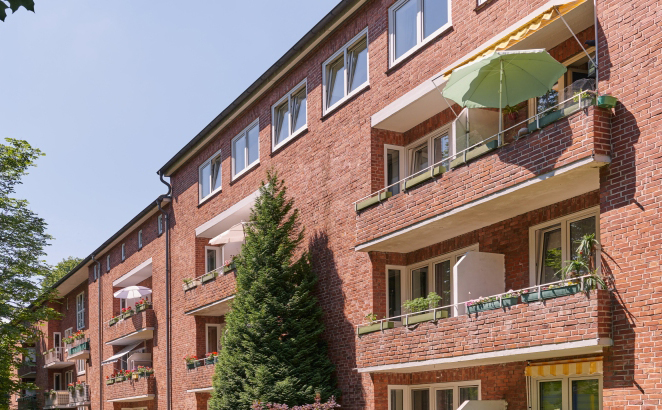 Ein Wohnhaus aus rotem Backstein mit Balkonen, teils mit Sonnenschirmen. ?berall hingetupft stehen unter blauem Himmel gr?ne B?ume und Pflanzen.