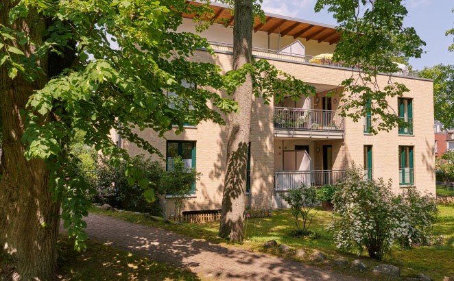 Ein modernes Mehrfamilienhaus mit heller Klinkerfassade und großzügigen Balkonen. Es befindet sich in einer gepflegten Grünanlage mit üppigen Bäumen.
