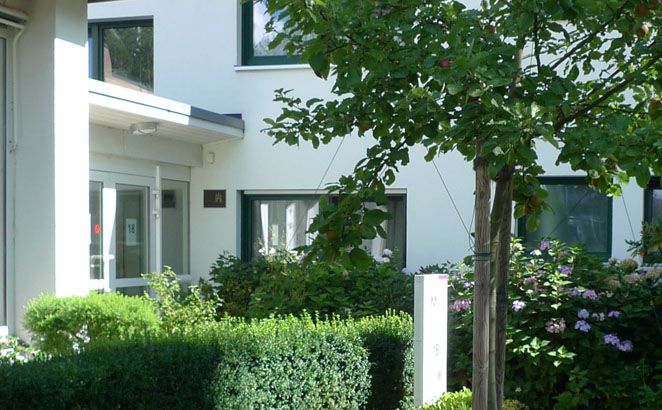 Der Eingang eines Wohngebäudes mit heller Fassade in einer grünen Umgebung mit gepflegten Büschen und Bäumen.