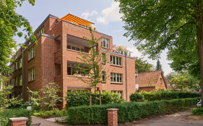 Mehrfamilienhaus mit roter Klinkerfassade und großen Balkonen in ruhiger Nachbarschaft. Es ist umgeben von Bäumen und gepflegten Hecken.